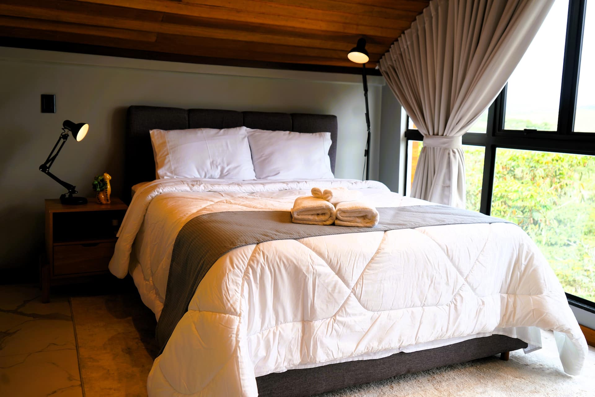 Cama de casal com toalhas dobradas, teto em tábuas de madeira e luminária de leitura