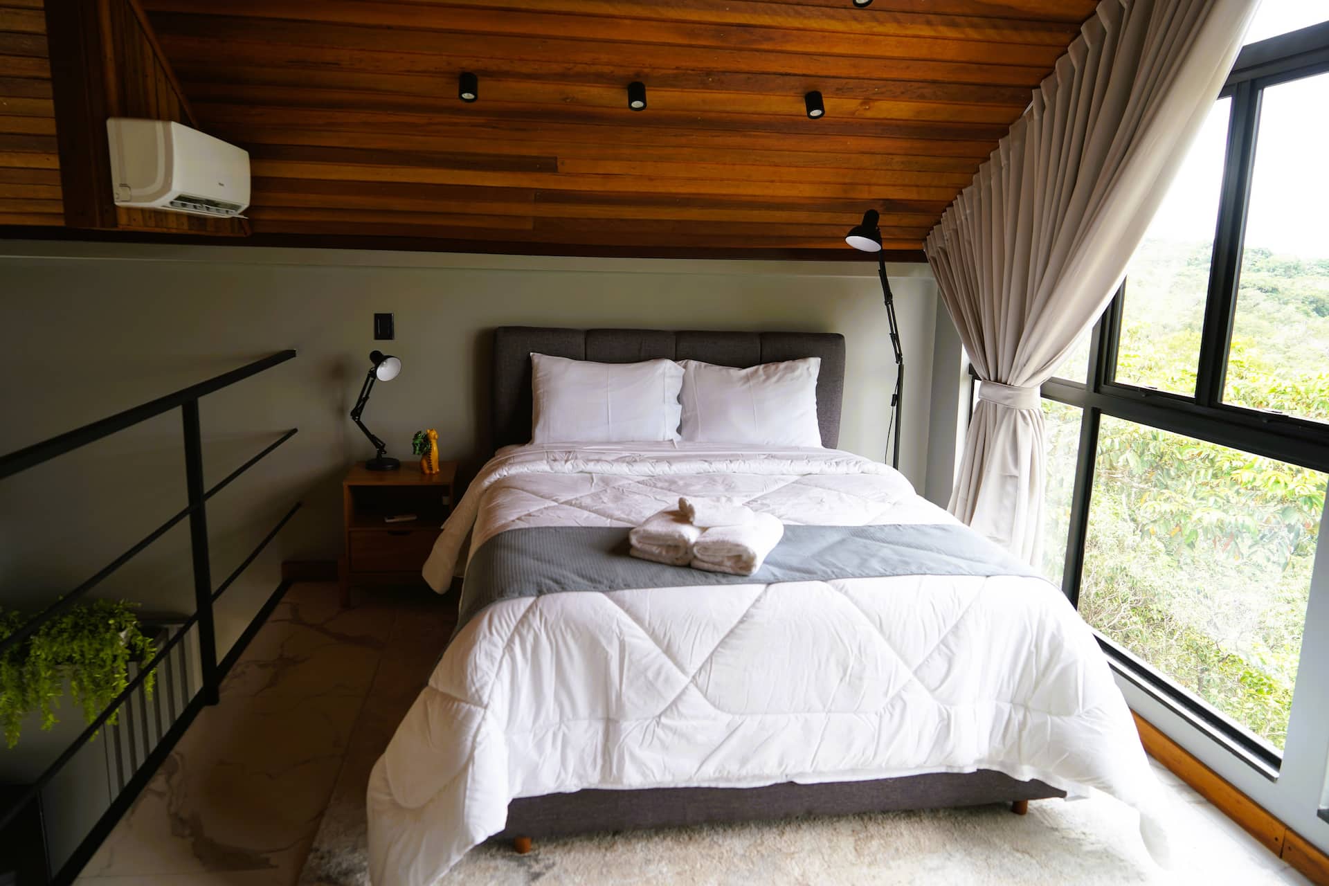 Cama vista do mezanino, com ar-condicionado, luminária articulada e janela para a mata
