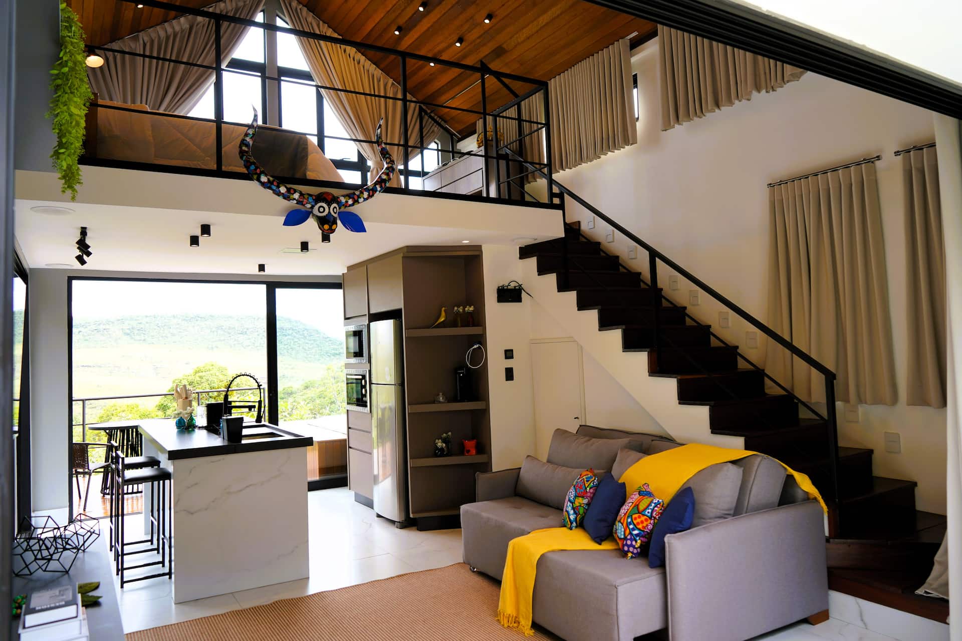Sala com pé-direito duplo, mezanino do quarto, escada preta e vista da serra pela vidraça